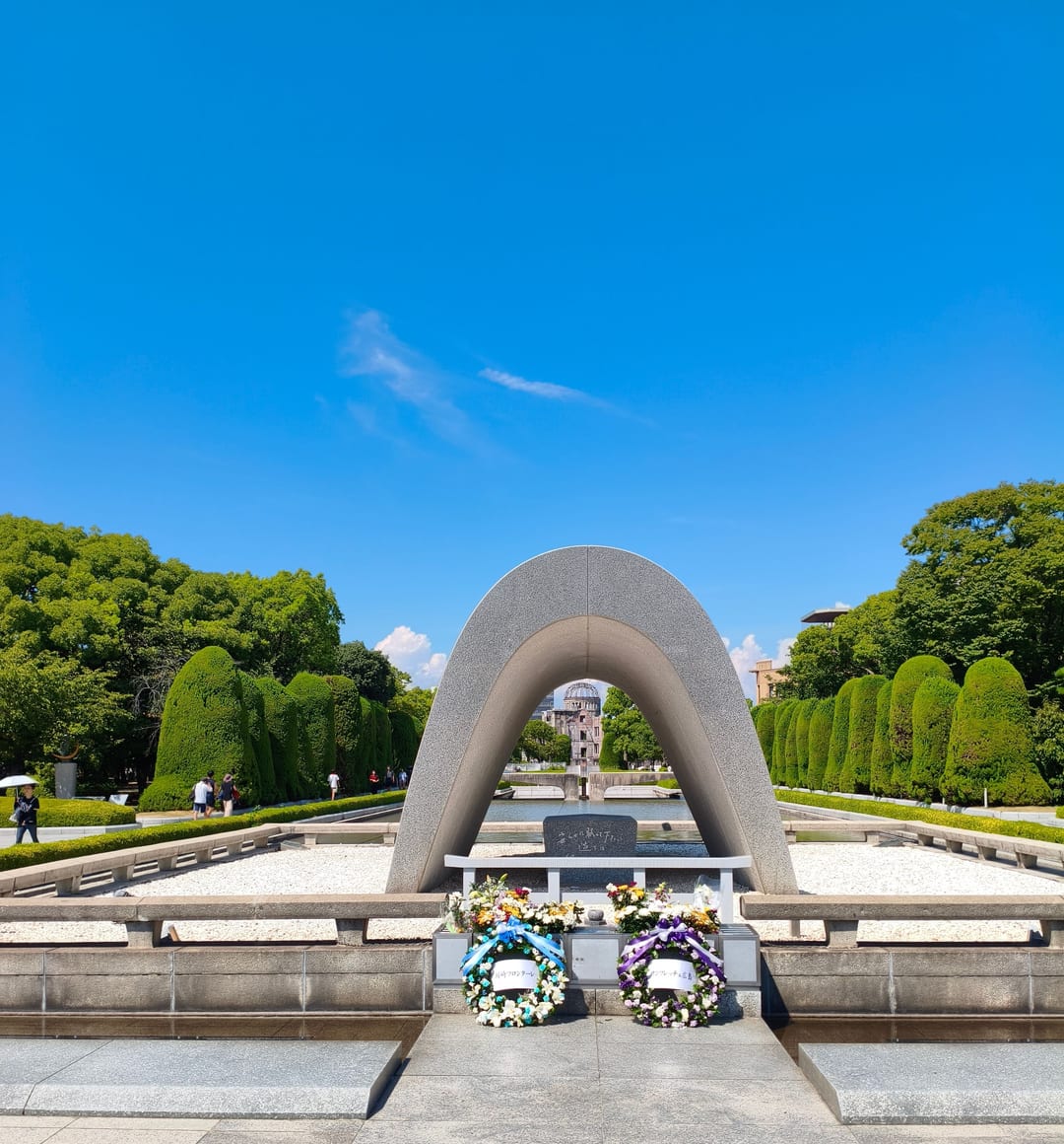 The arched concrete Cenotaph with wreaths laid in front of it, the Atomic Bomb Dome visible in the distance through the arch
