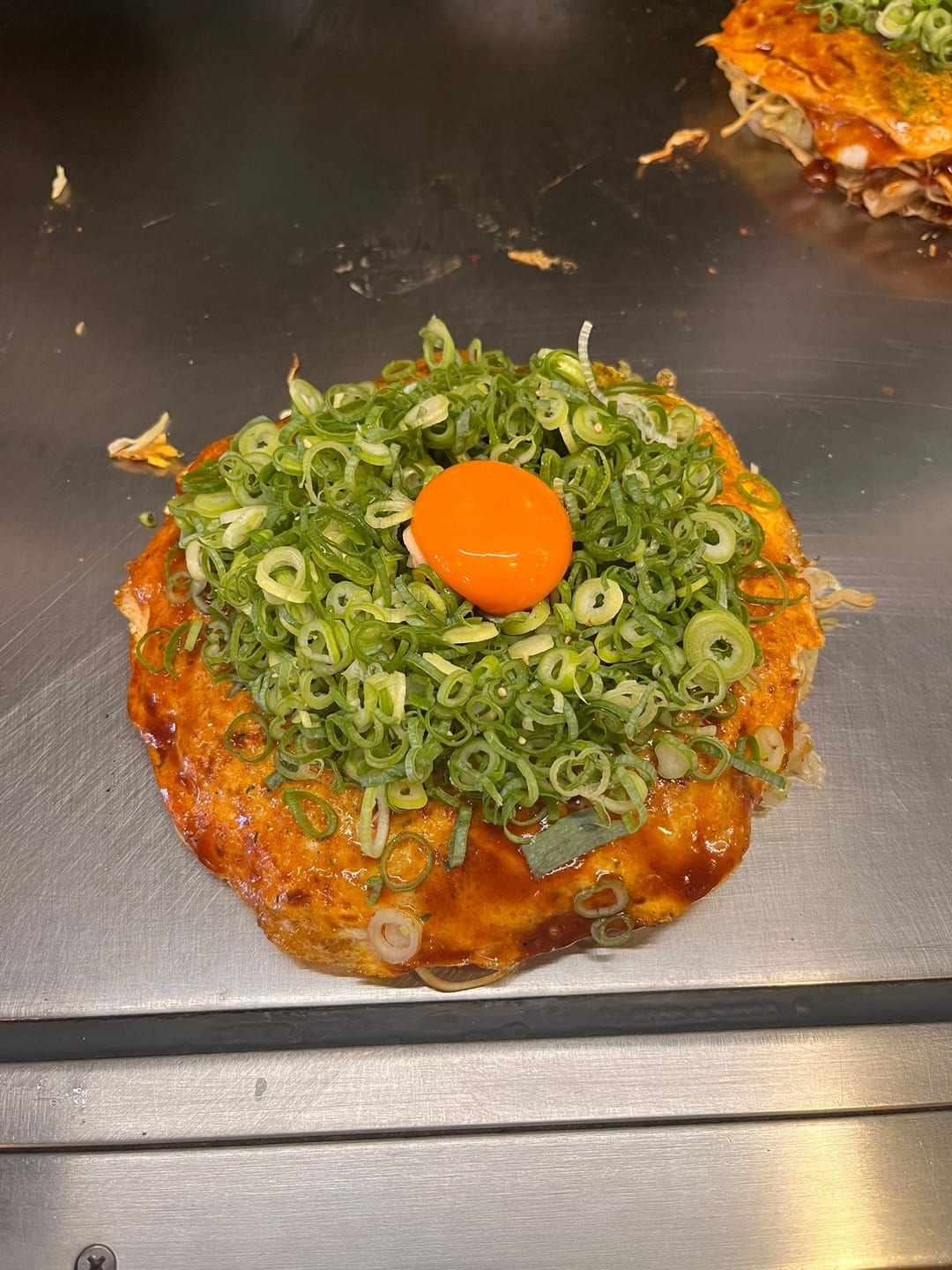 An okonomiyaki cooking on a hot griddle, topped with an egg yolk and a ring of chopped spring onion