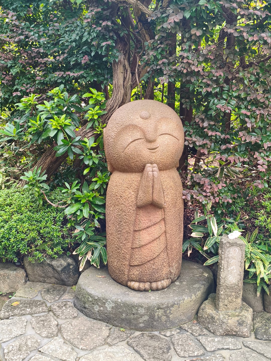 A small stone statue of a monk with hands pressed together in prayer, surrounded by greenery