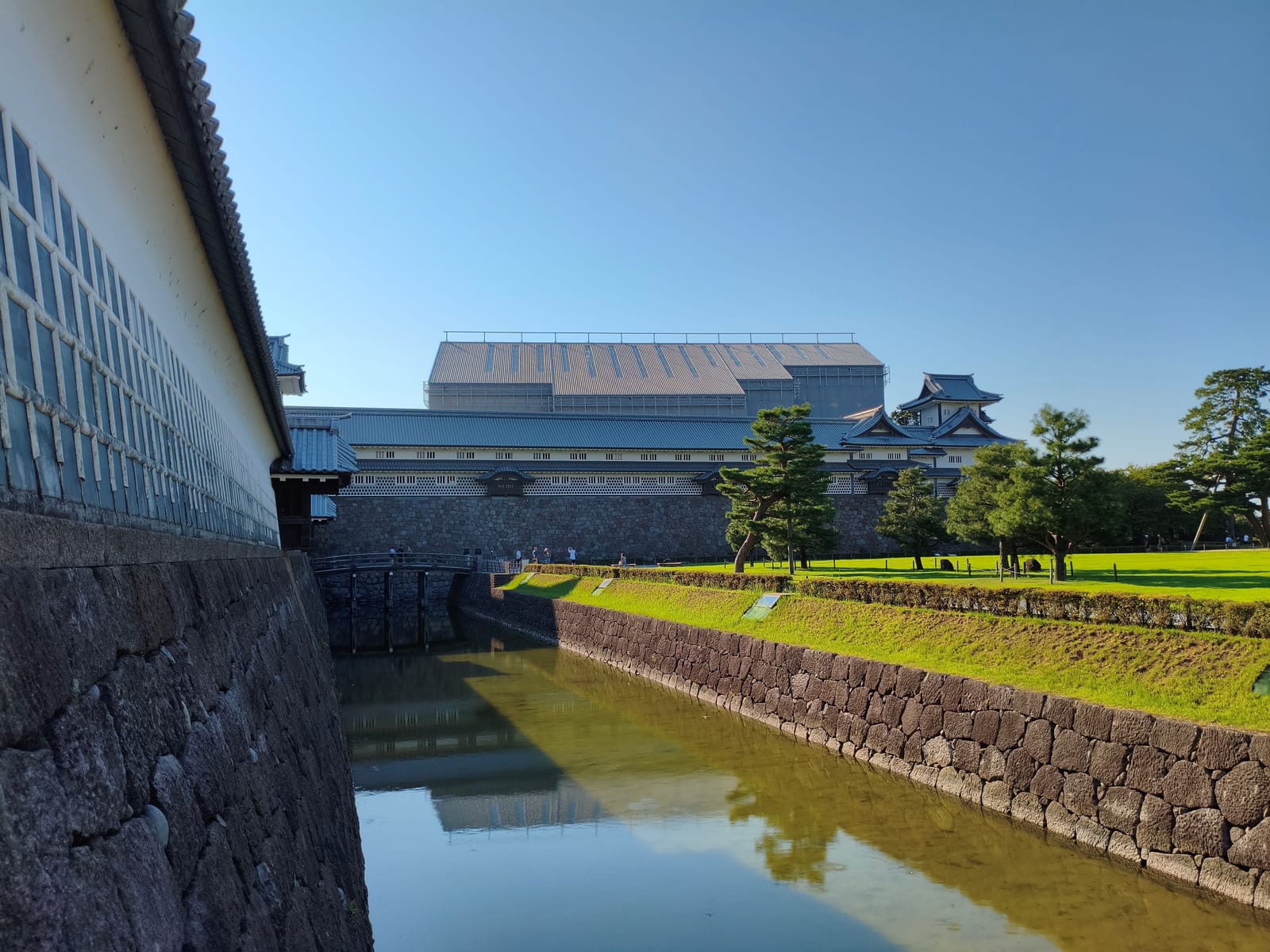 The white walls and stone base of Kanazawa Castle reflected in its moat under a clear sky