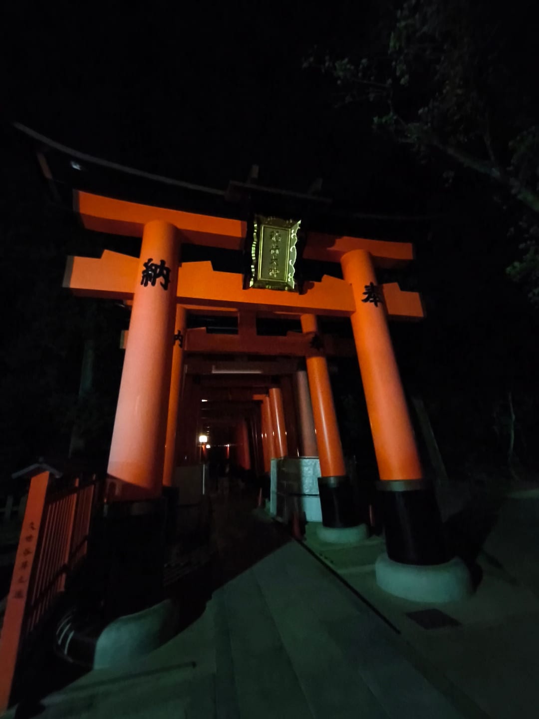 The entrance to the Senbon Torii at night, an illuminated plaque above the gate and the tunnel of torii running away into the dark