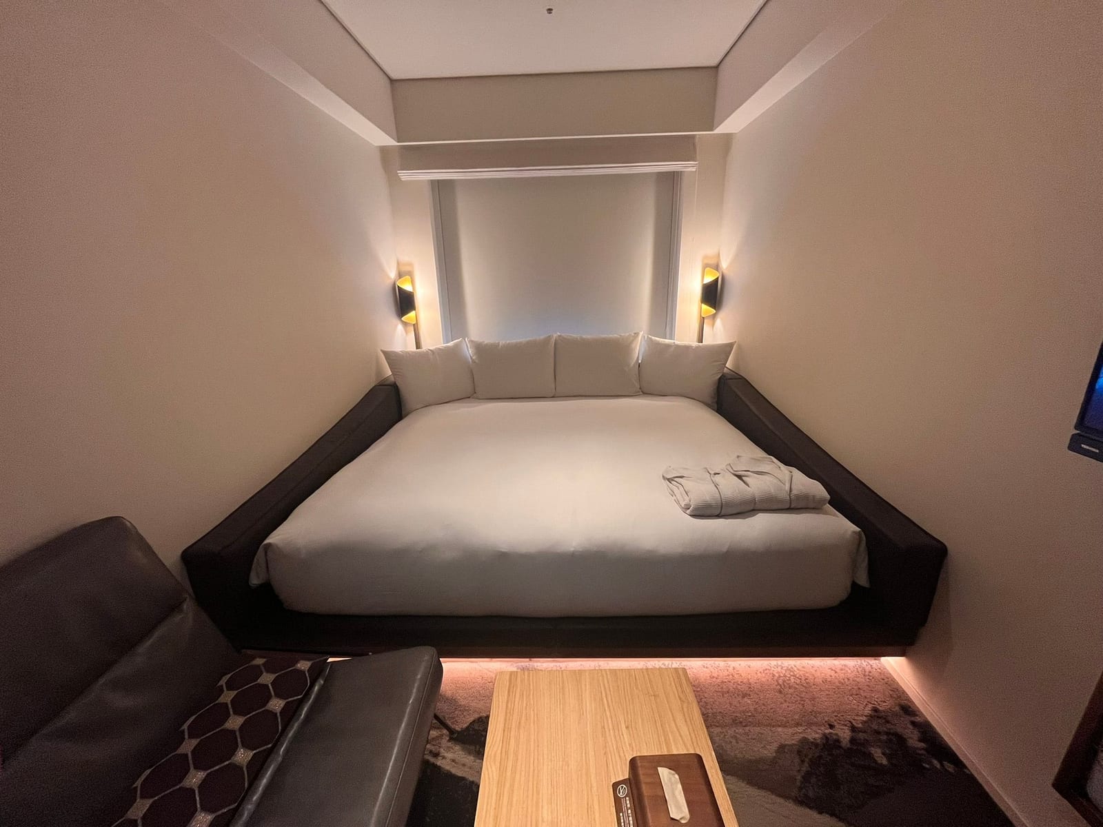 A hotel room with a large bed set into an alcove between two walls, lit by wall lamps, with a sofa and a low table in the foreground
