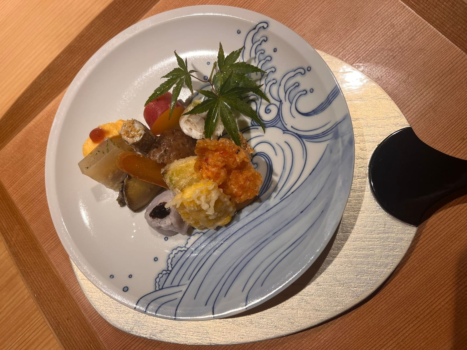 A kaiseki appetiser course arranged on a plate painted with a blue wave, garnished with a maple leaf