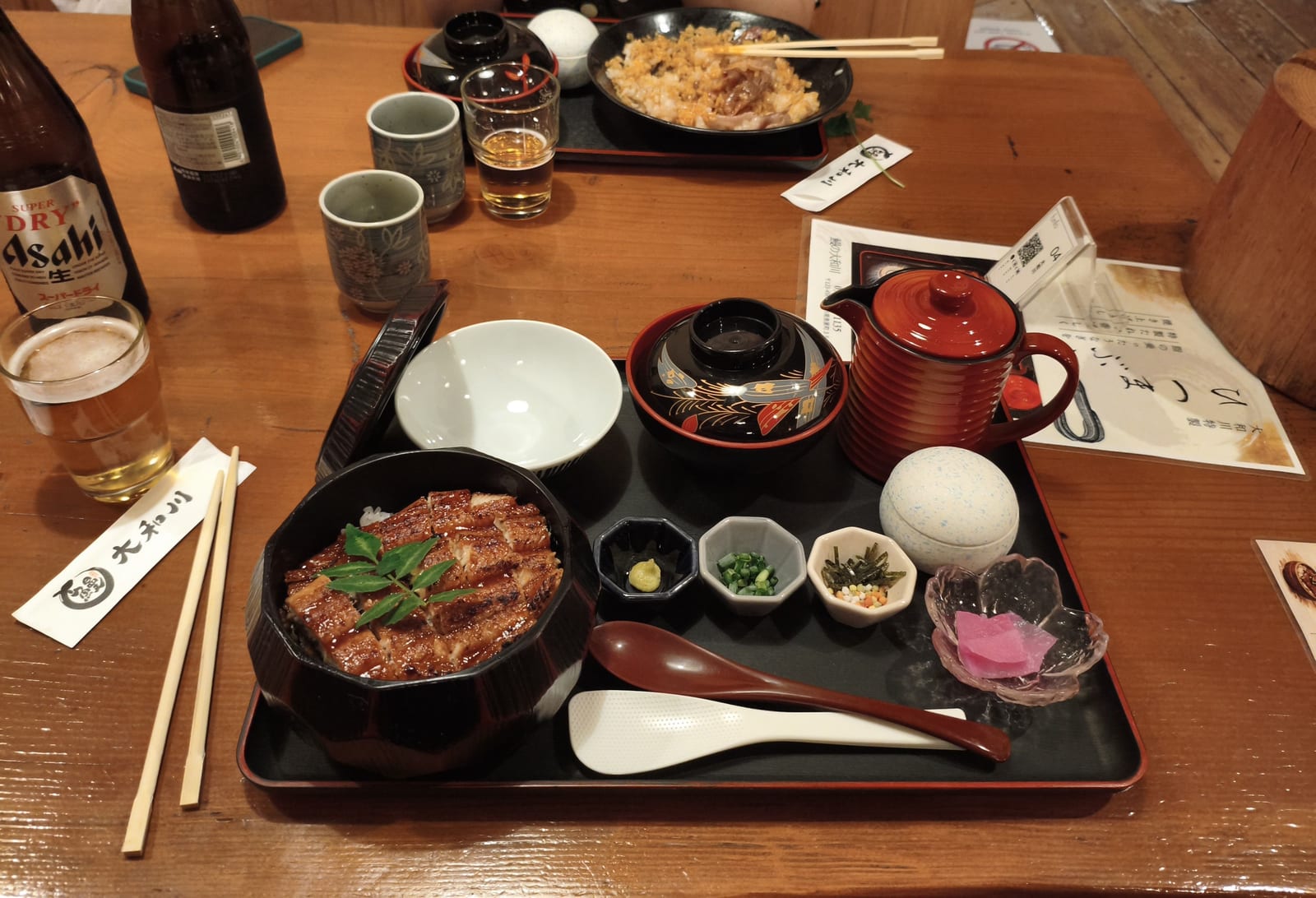 A tray holding glazed eel over rice in a lacquer bowl, with pickles, side dishes and a pot of broth