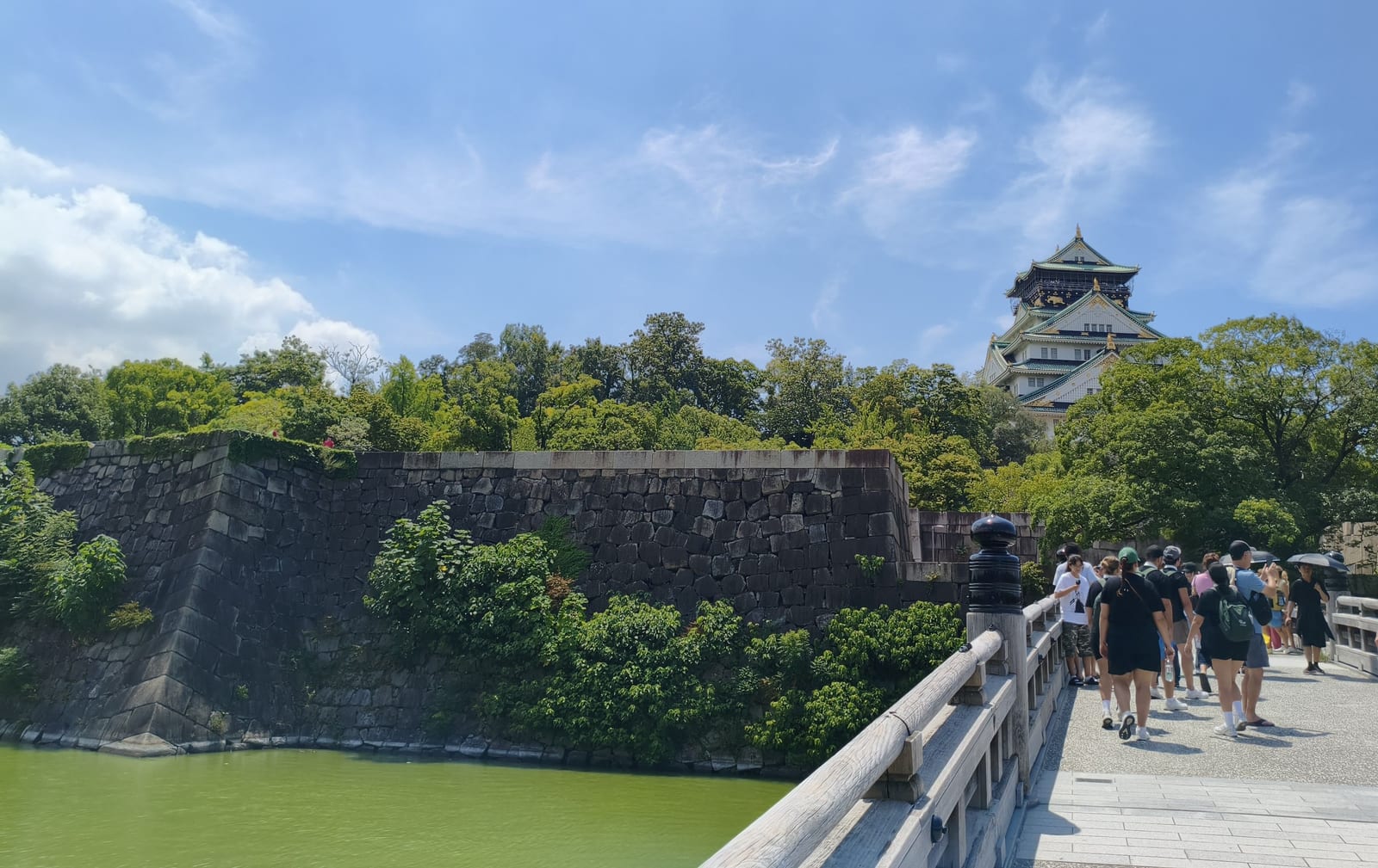 The moat and huge sloping stone walls of Osaka Castle, with the keep rising behind the trees and visitors crossing a bridge