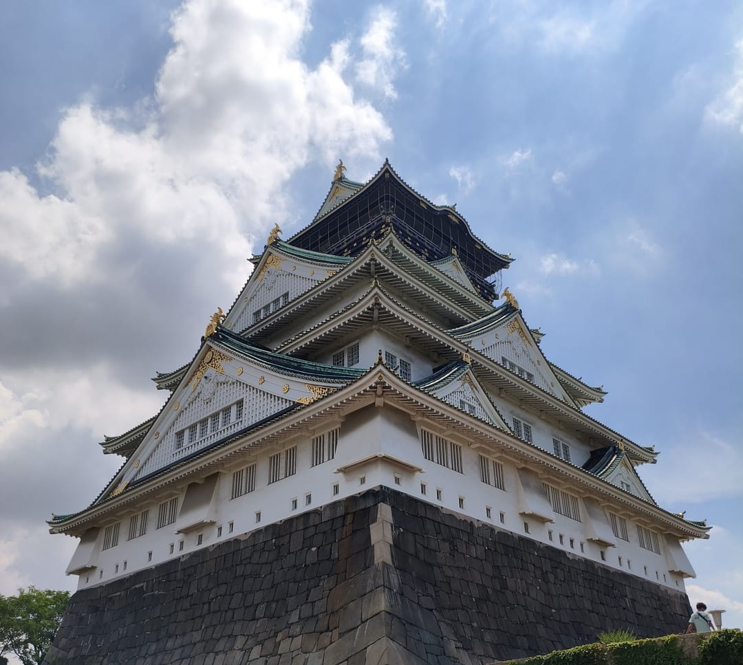 The main keep of Osaka Castle seen from below, its white tiers trimmed with green and gold against a cloudy sky