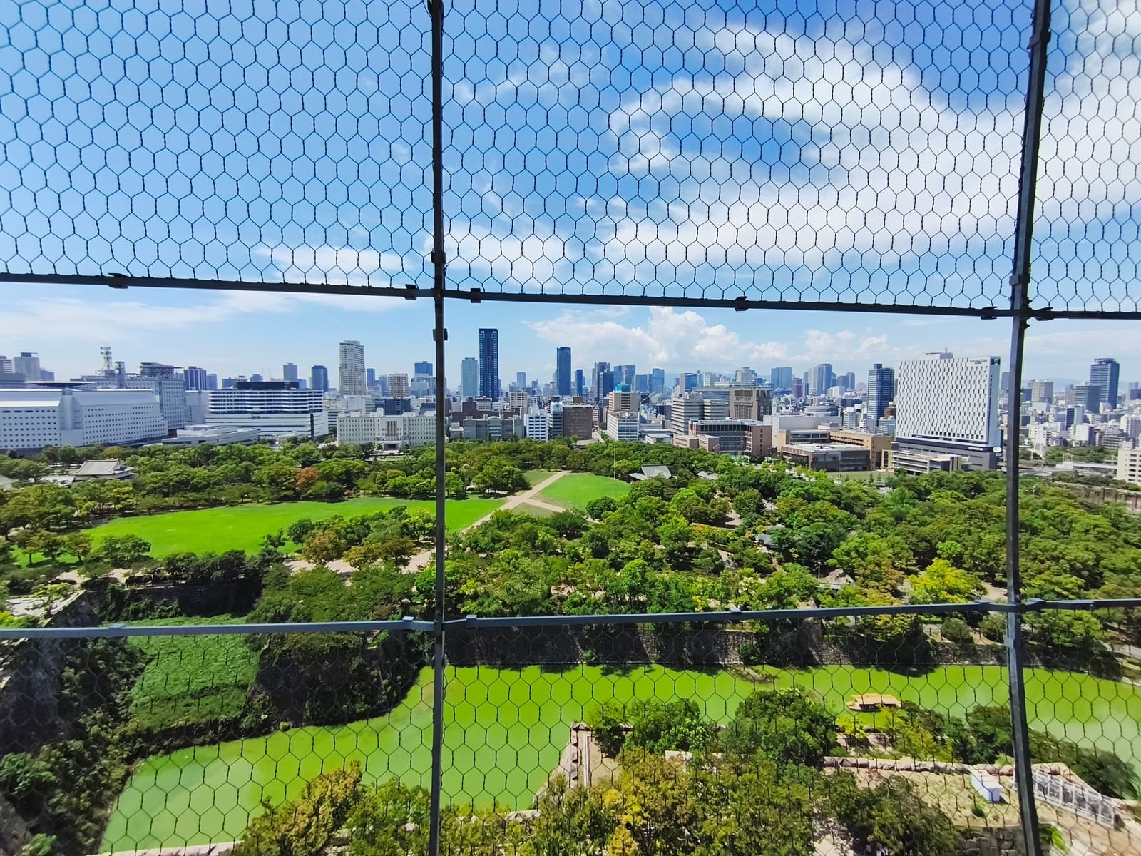 The view from the castle observation deck over the park, the moat and the Osaka skyline, seen through safety mesh