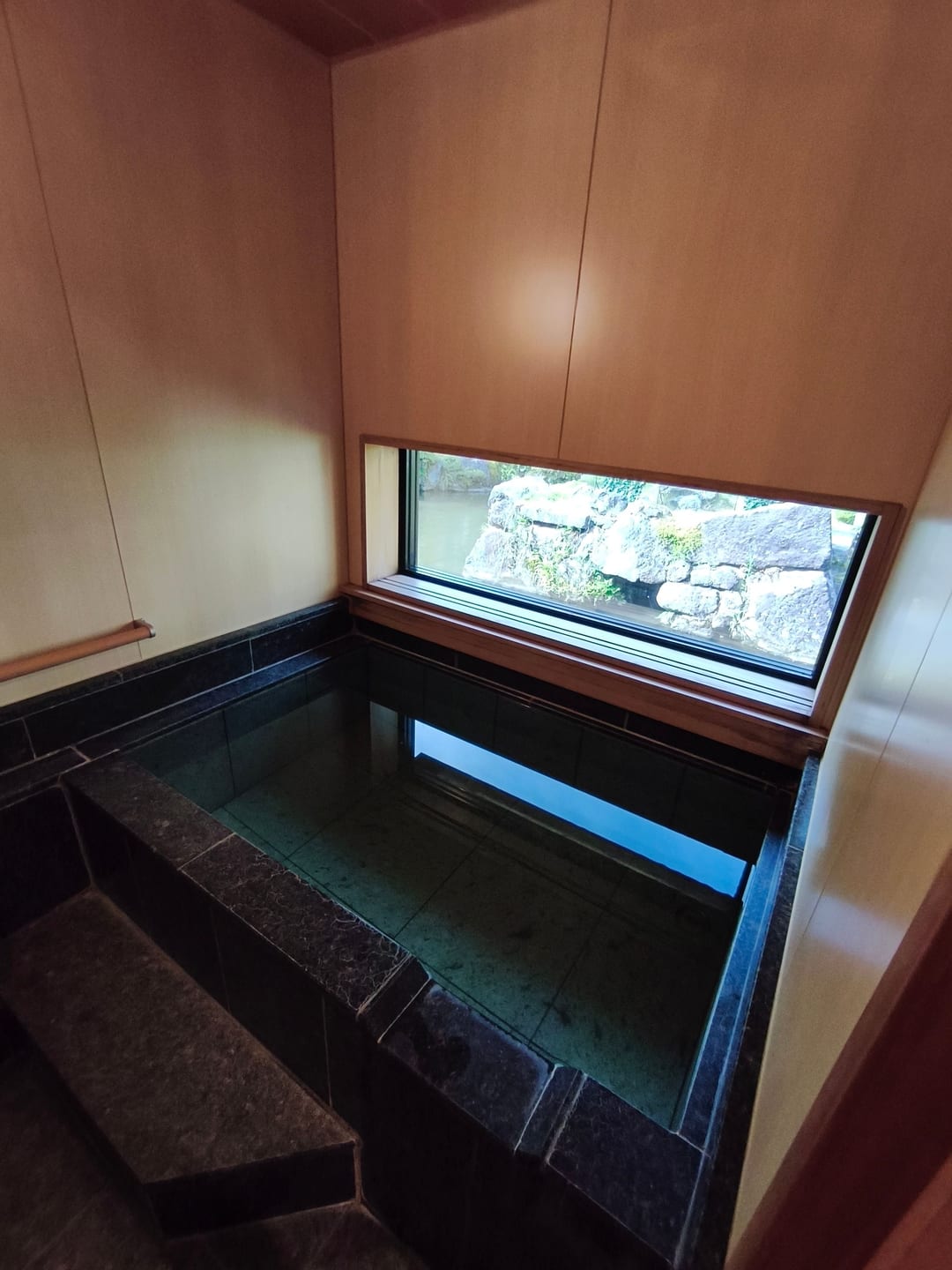 A deep stone bath in a wood-panelled room, with a long narrow window looking out onto greenery