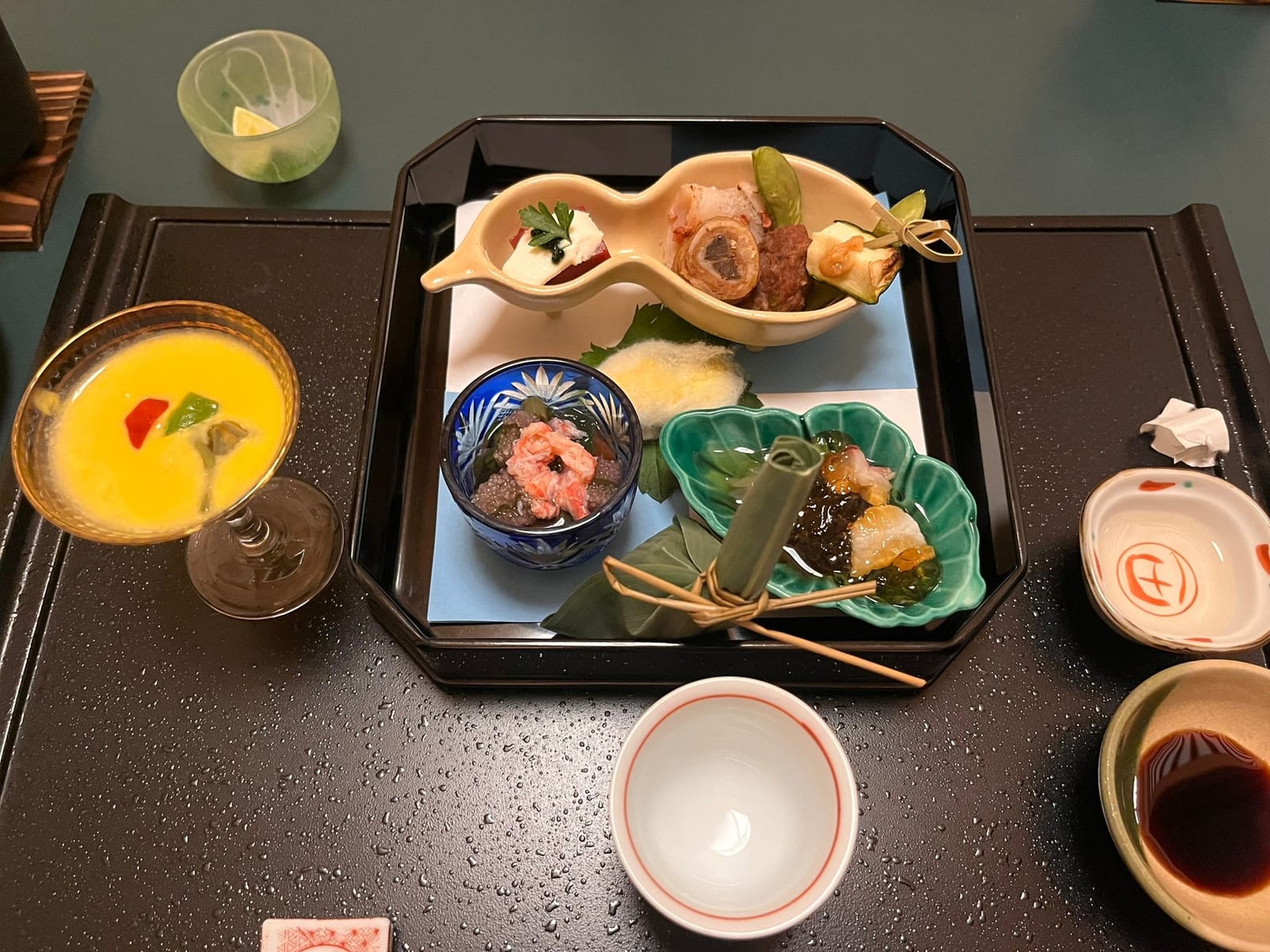 A lacquer tray of small kaiseki dishes with a chilled drink and sauces set out on a dark table