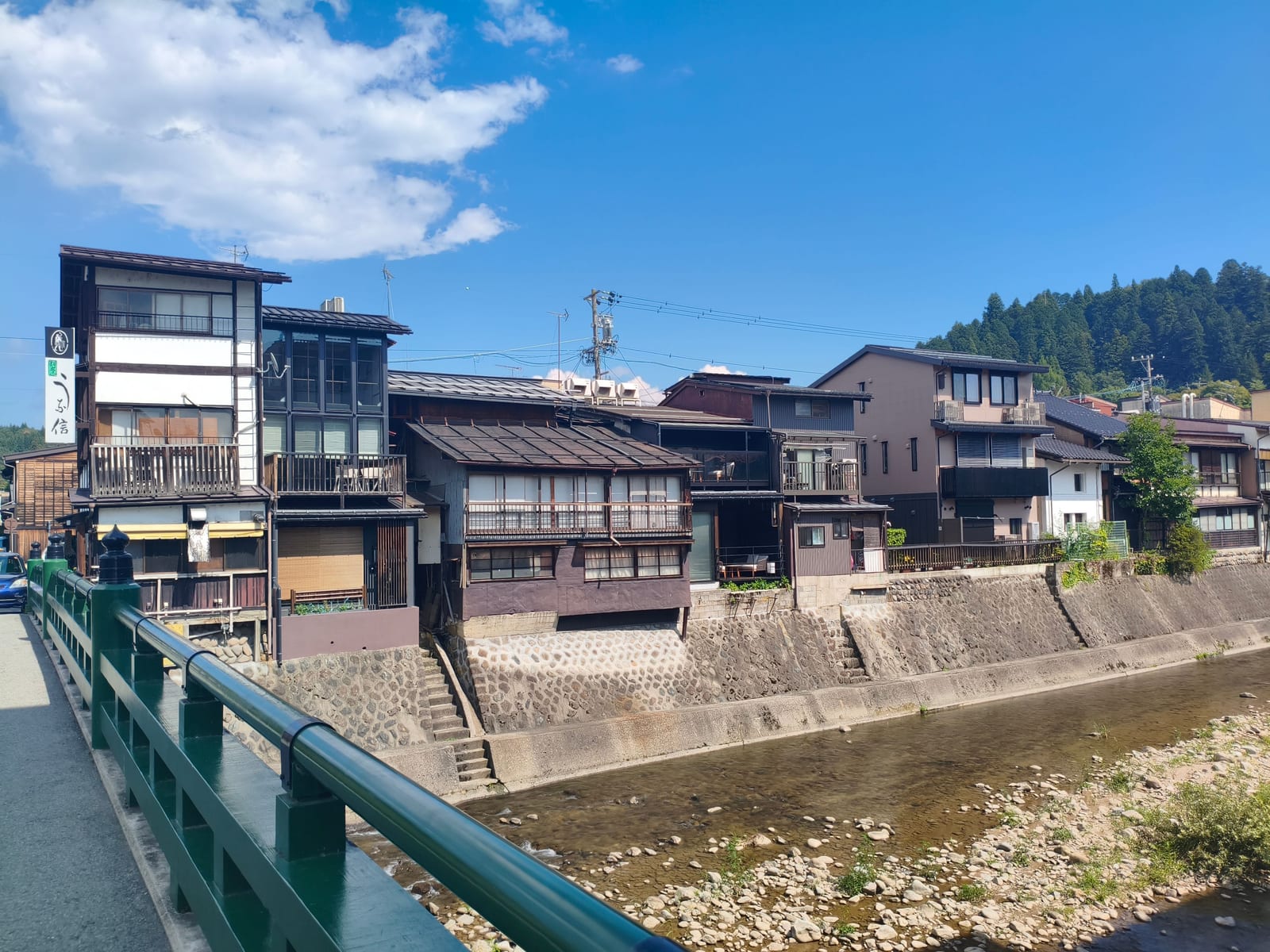 Traditional houses lining the stone embankment of the river through Takayama, a green bridge railing in front
