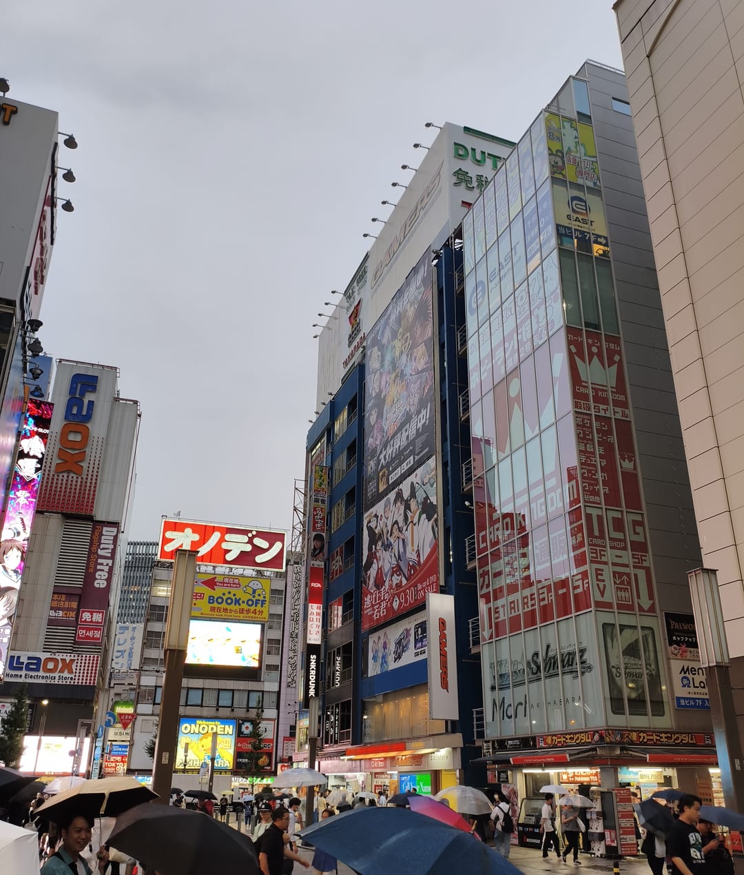 A street in Akihabara under a grey sky, signage stacked up the buildings and people with umbrellas below